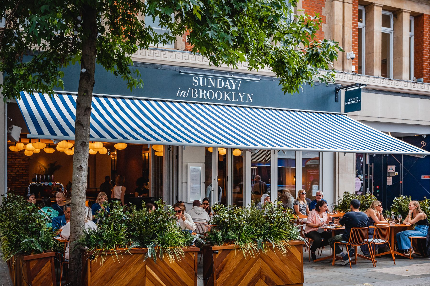 Sunday in Brooklyn, James Street. Shopfront with striped awning, signage, and pavement dining.